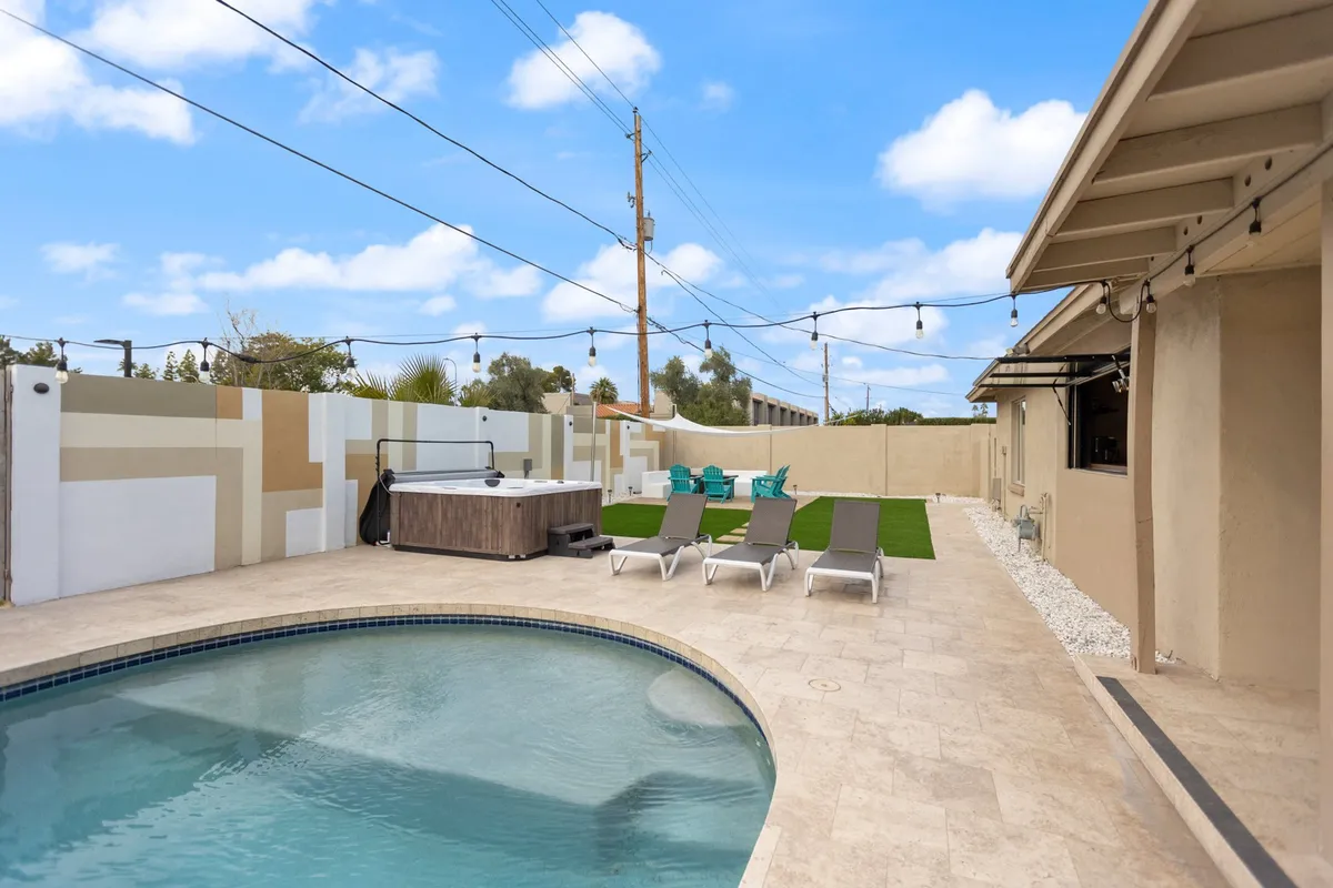 This vibrant exterior scene showcases a sparkling swimming pool, an inviting hot tub, several lounge chairs, a well-manicured green lawn, and a stylish decorative wall. The layout suggests a space perfect for sunbathing, relaxing in the hot tub, poolside gatherings, and enjoying the outdoors with friends and family.
