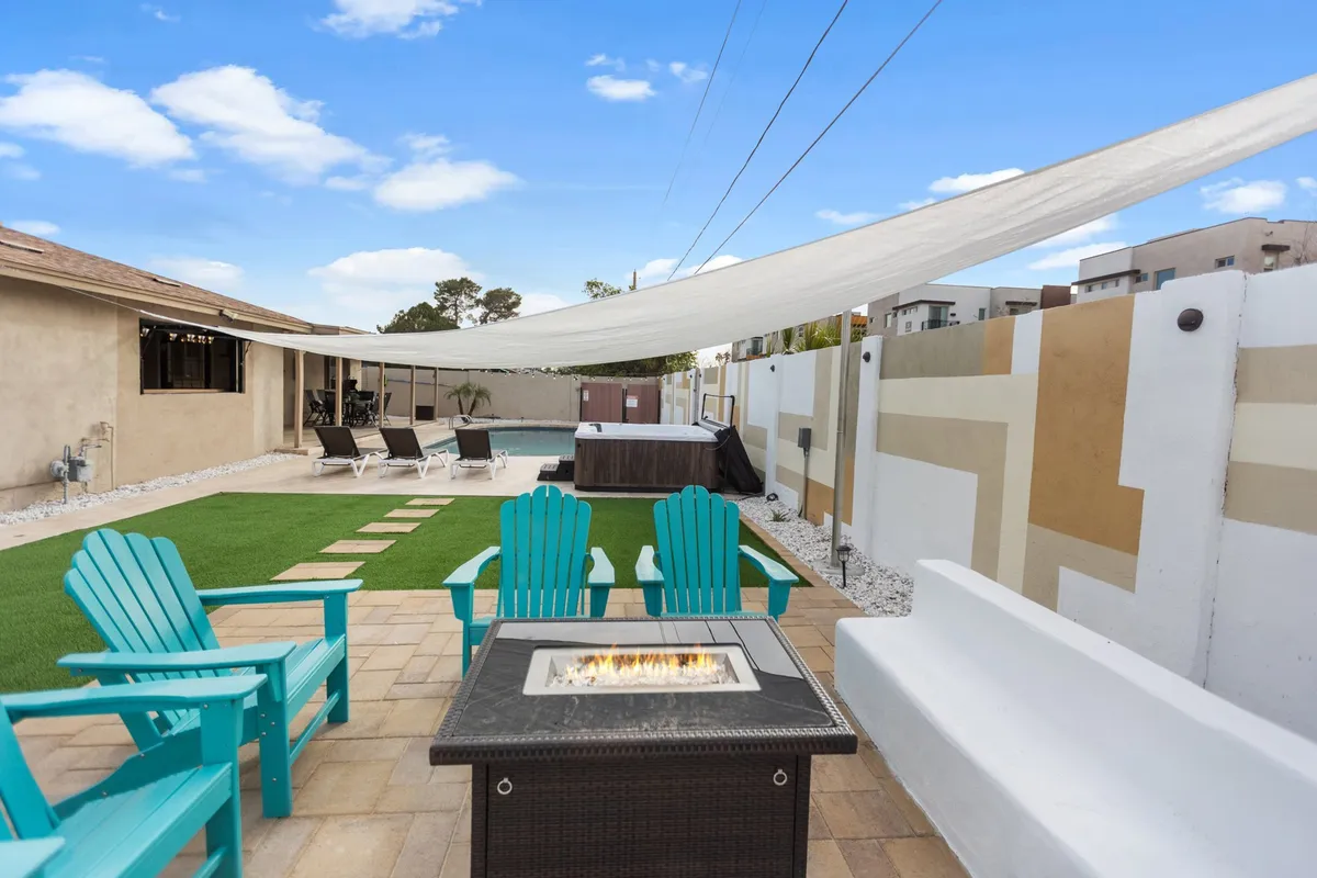 An inviting backyard scene with a fire pit surrounded by Adirondack chairs, a lawn area, pool, and a shade sail. The space is designed for both daytime relaxation and evening gatherings, with a strong sense of social connection and comfort.