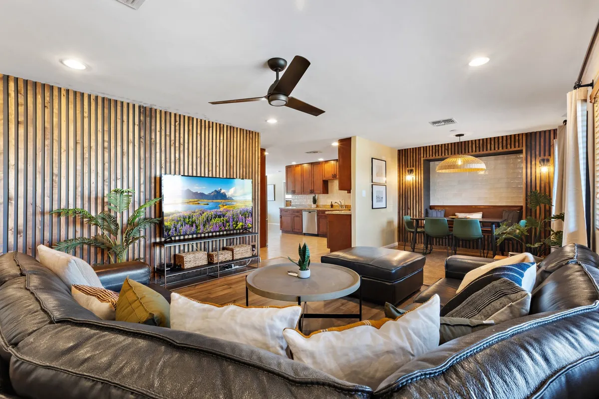 This image features a spacious open-concept living and dining area. A large sectional sofa faces a TV set against a wood panel accent wall, ideal for relaxing or group entertainment. In the background, the dining area is visible, illustrating the seamless flow between spaces and highlighting the communal atmosphere.
