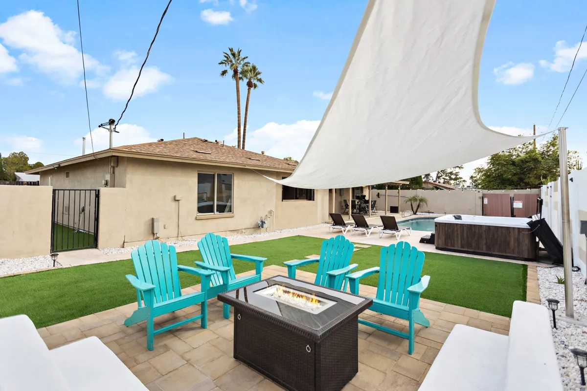 A vibrant backyard scene featuring a fire pit with Adirondack chairs, a sparkling swimming pool, shade sail, and palm trees. The space is ideal for group gatherings, evening conversations, and enjoying the Scottsdale climate after exploring town.