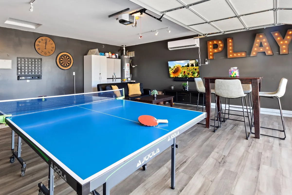 This image showcases a vibrant game room complete with a ping pong table, a playful 'PLAY' sign on the wall, a large TV displaying a nature scene, dartboard, and a clock. The space is bright and energetic, with clear zones for both active games and relaxed entertainment.
