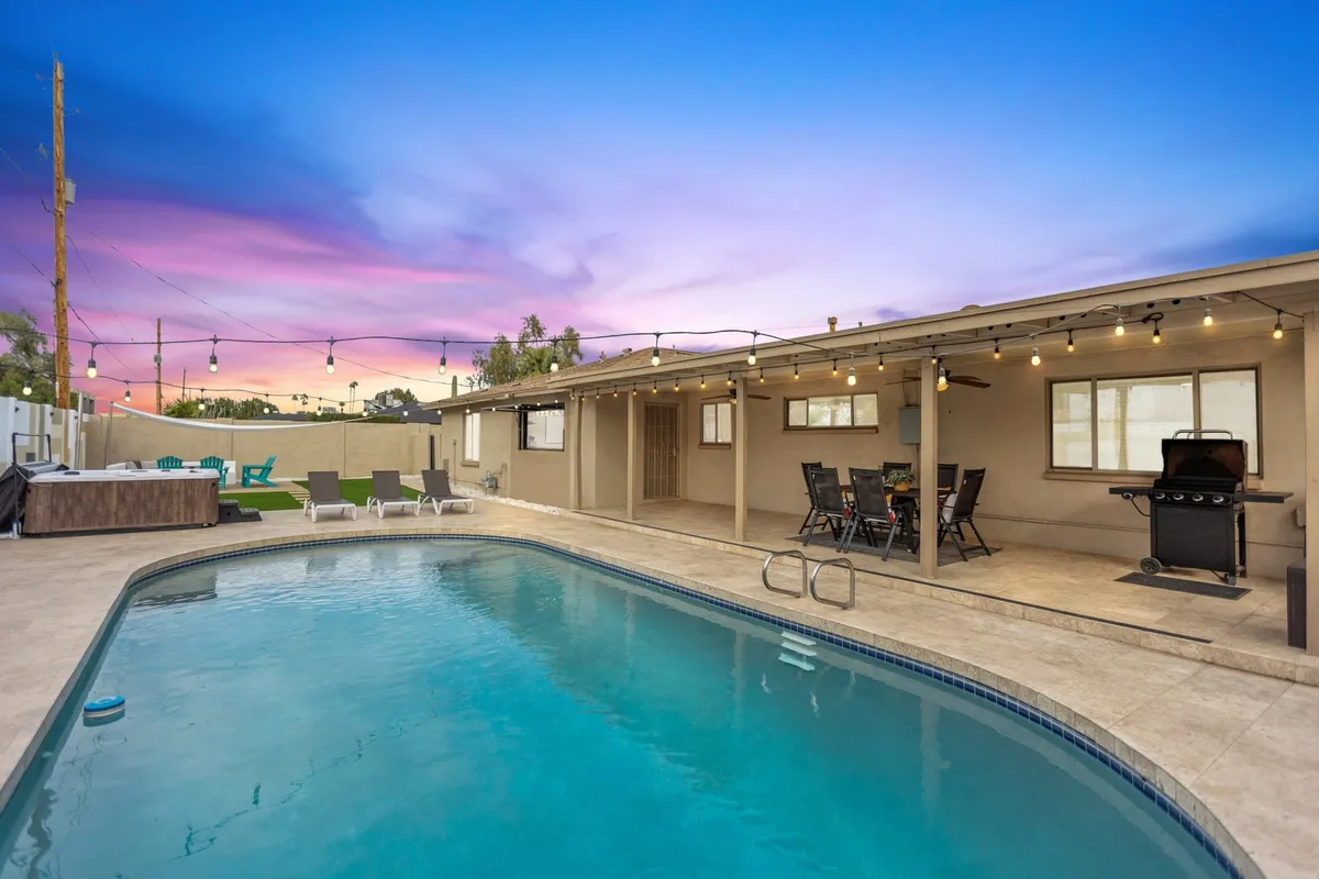 The scene centers on a sparkling pool flanked by lounge chairs, an outdoor seating area with a grill, and festive string lights overhead. Sunset colors paint the sky, creating a magical ambiance for evening gatherings. The area is set up for barbecuing, relaxing, and entertaining groups outdoors.