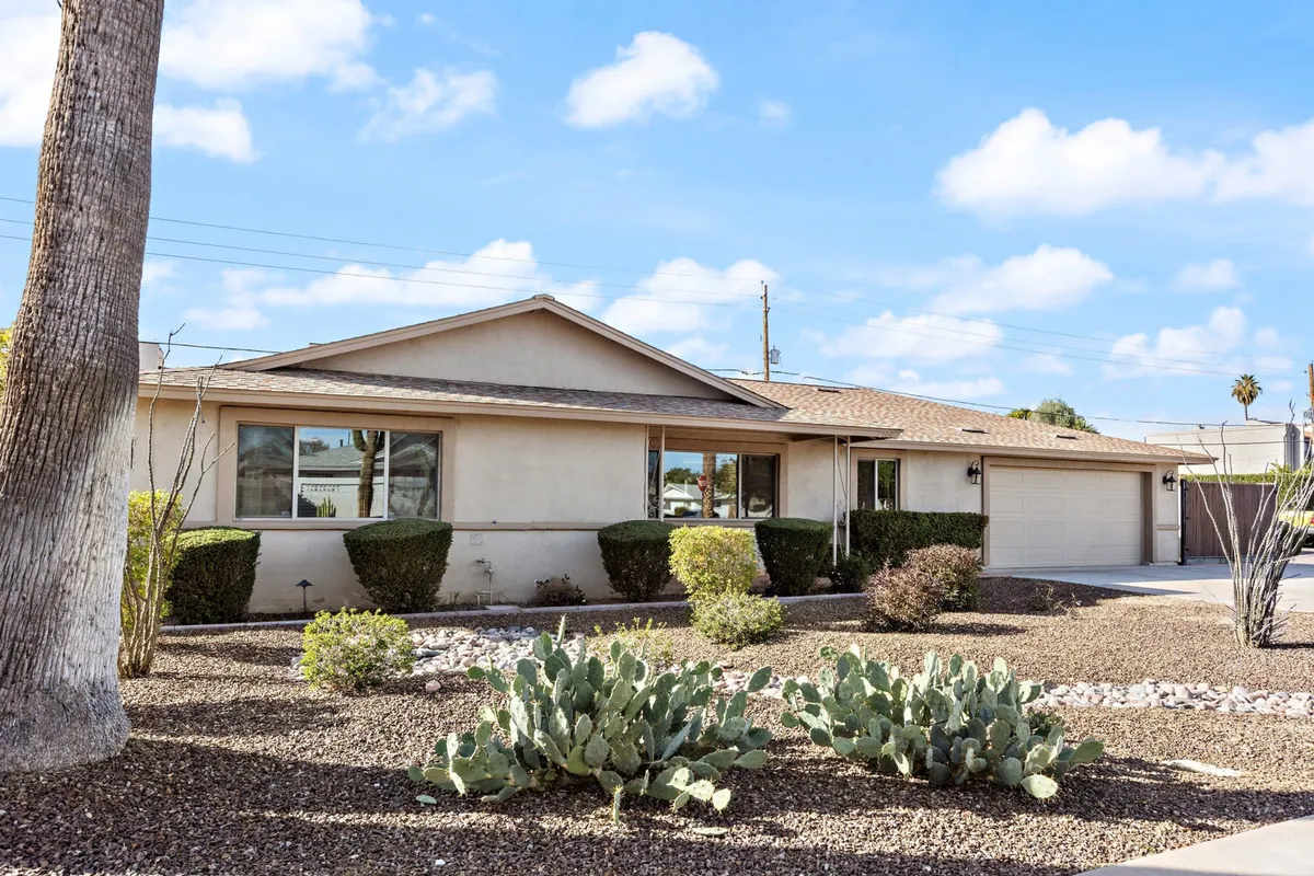 The image showcases the home's inviting front garden, adorned with classic Arizona cacti, tall palm trees, and a warm facade. The scene radiates the welcoming atmosphere of a Scottsdale neighborhood and hints at the proximity to lively city attractions.