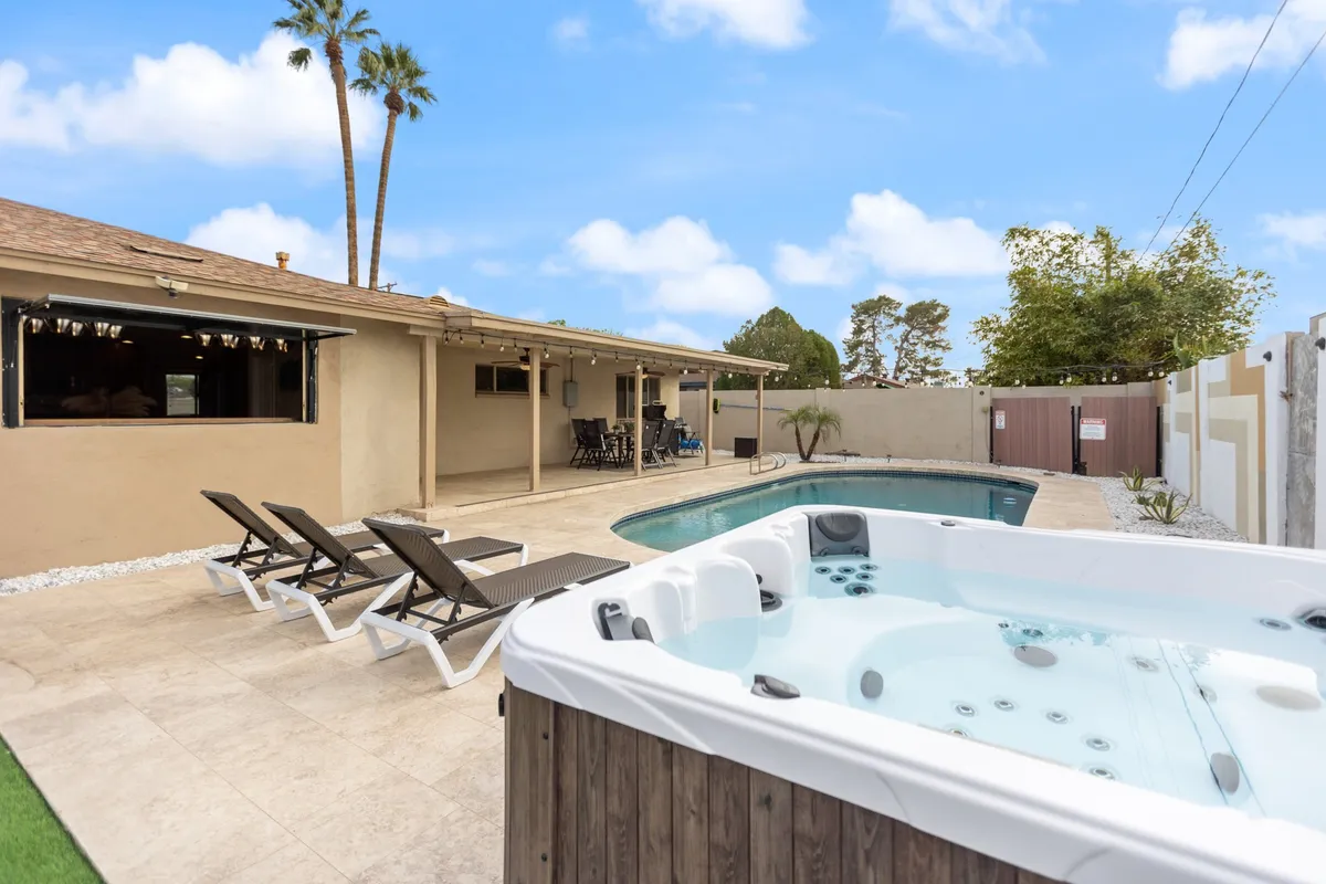 A beautifully landscaped backyard featuring a sparkling swimming pool and adjacent hot tub. Lounge chairs surround the water, while a covered patio and seating area provide ample space for guests to relax or entertain outdoors. The sun casts a warm glow, creating an inviting and tranquil resort-style atmosphere.